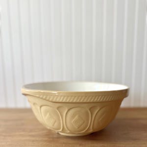 K&ouml;nnte beinhalten: Eine beige Keramik-R&uuml;hrsch&uuml;ssel mit cremefarbenem Innenraum. Die Sch&uuml;ssel hat ein dekoratives Rautenmuster und einen strukturierten Rand. Sie steht auf einer Holzoberfl&auml;che vor einem wei&szlig;en Hintergrund.