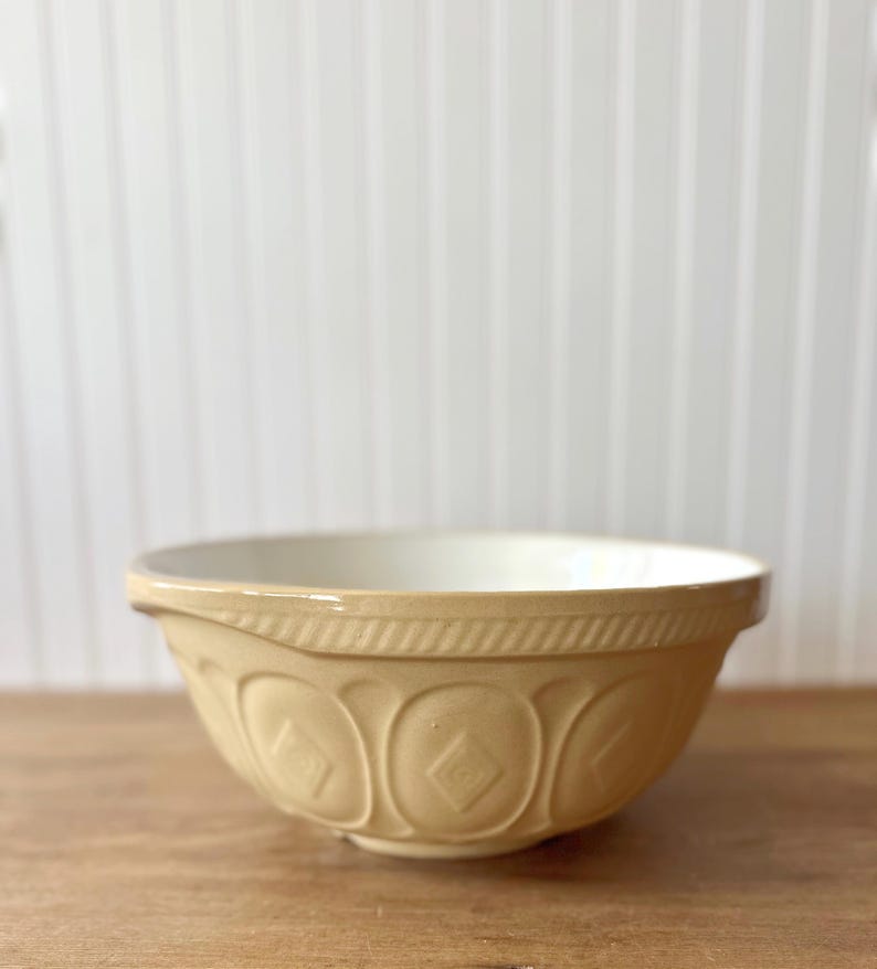 K&ouml;nnte beinhalten: Eine beige Keramik-R&uuml;hrsch&uuml;ssel mit cremefarbenem Innenraum. Die Sch&uuml;ssel hat ein dekoratives Rautenmuster und einen strukturierten Rand. Sie steht auf einer Holzoberfl&auml;che vor einem wei&szlig;en Hintergrund.
