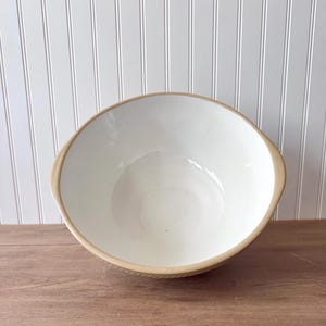 K&ouml;nnte beinhalten: Eine gro&szlig;e, cremefarbene Keramik-R&uuml;hrsch&uuml;ssel mit einem beigefarbenen Rand und &Auml;u&szlig;eren. Die Sch&uuml;ssel hat ein breites, offenes Design und steht auf einer Holzoberfl&auml;che. Der Innenraum ist glatt und das &Auml;u&szlig;ere hat ein strukturiertes Muster.