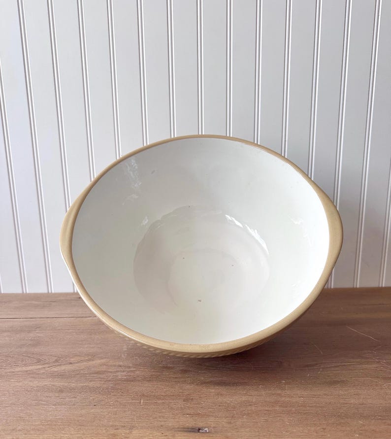 K&ouml;nnte beinhalten: Eine gro&szlig;e, cremefarbene Keramik-R&uuml;hrsch&uuml;ssel mit einem beigefarbenen Rand und &Auml;u&szlig;eren. Die Sch&uuml;ssel hat ein breites, offenes Design und steht auf einer Holzoberfl&auml;che. Der Innenraum ist glatt und das &Auml;u&szlig;ere hat ein strukturiertes Muster.