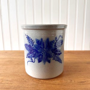 Puede incluir: Un tarro de gres con un diseño floral azul. El recipiente cilíndrico tiene un acabado moteado gris claro y un motivo de flor y hoja azul oscuro. El tarro está sobre una superficie de madera.