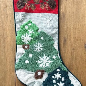 Puede incluir: Un calcetín navideño con la parte superior roja con piñas y ramitas. El cuerpo es blanco con árboles de Navidad verdes y copos de nieve. Tiene un ribete verde y un lazo para colgar.
