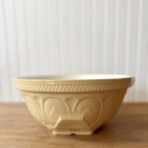 K&ouml;nnte beinhalten: Eine beige Keramik-R&uuml;hrsch&uuml;ssel mit wei&szlig;em Innenraum. Die Sch&uuml;ssel hat ein dekoratives gepr&auml;gtes Muster auf der Au&szlig;enseite und einen breiten Rand. Die Sch&uuml;ssel steht auf einer Holzoberfl&auml;che.