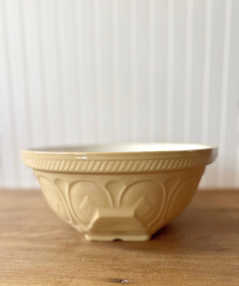 K&ouml;nnte beinhalten: Eine beige Keramik-R&uuml;hrsch&uuml;ssel mit wei&szlig;em Innenraum. Die Sch&uuml;ssel hat ein dekoratives gepr&auml;gtes Muster auf der Au&szlig;enseite und einen breiten Rand. Die Sch&uuml;ssel steht auf einer Holzoberfl&auml;che.