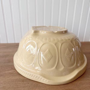 K&ouml;nnte beinhalten: Eine cremefarbene, vintage Keramik-R&uuml;hrsch&uuml;ssel. Die Sch&uuml;ssel hat ein dekoratives Muster aus ovalen Formen mit Rautenmustern. Die Sch&uuml;ssel steht verkehrt herum auf einer Holzoberfl&auml;che.