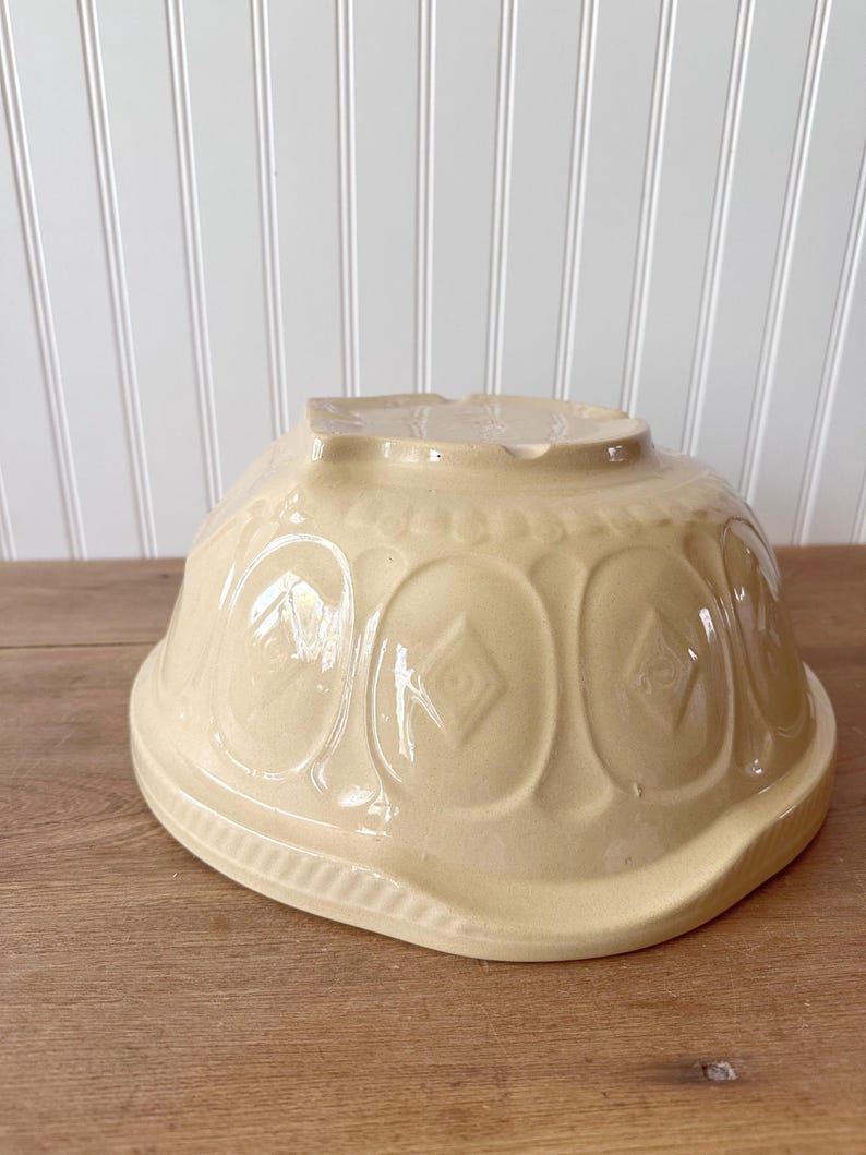 K&ouml;nnte beinhalten: Eine cremefarbene, vintage Keramik-R&uuml;hrsch&uuml;ssel. Die Sch&uuml;ssel hat ein dekoratives Muster aus ovalen Formen mit Rautenmustern. Die Sch&uuml;ssel steht verkehrt herum auf einer Holzoberfl&auml;che.