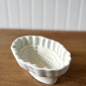 May include: A white ceramic corn-shaped mold with a scalloped edge. The mold is designed to stand upright and features detailed interior textures. The background is a wooden surface and a white wall.
