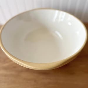 K&ouml;nnte beinhalten: Eine Keramiksch&uuml;ssel mit cremefarbenem Innenraum und beigem Rand. Die Sch&uuml;ssel steht auf einer Holzoberfl&auml;che. Das Design der Sch&uuml;ssel ist schlicht und klassisch, geeignet zum Servieren von Speisen oder als Dekoration.