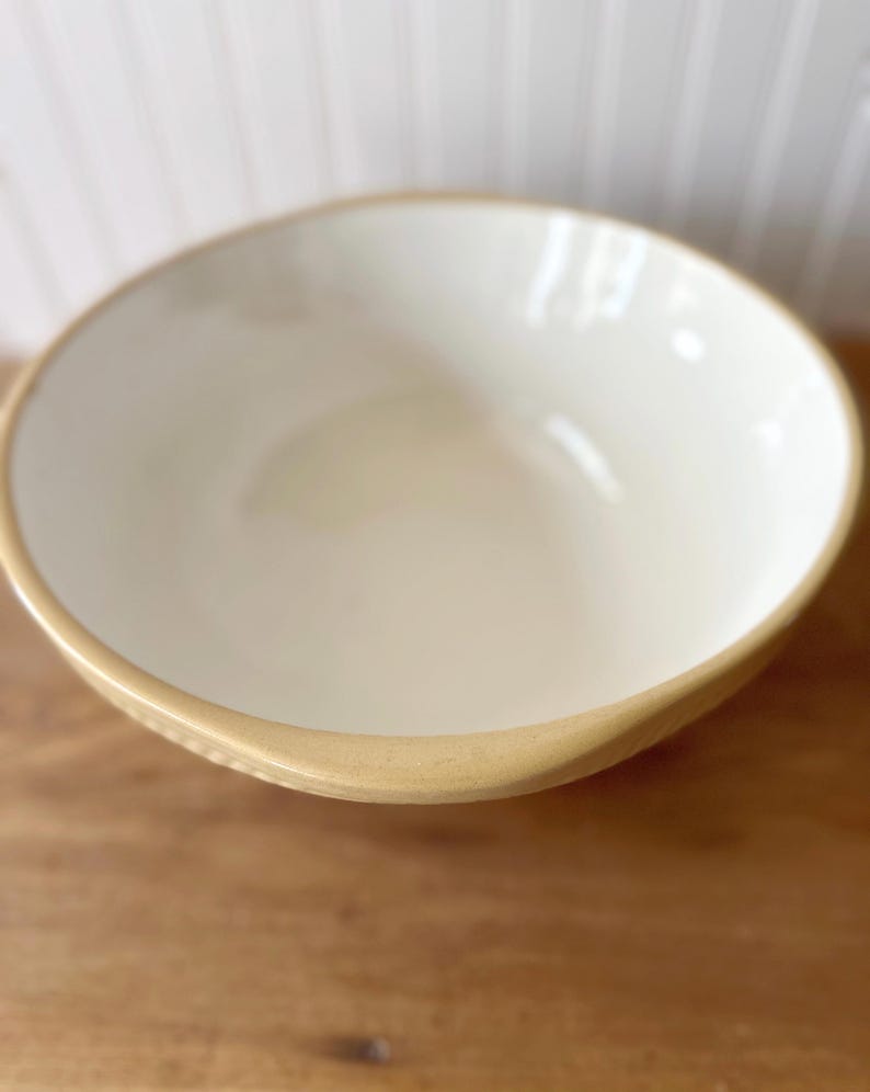 K&ouml;nnte beinhalten: Eine Keramiksch&uuml;ssel mit cremefarbenem Innenraum und beigem Rand. Die Sch&uuml;ssel steht auf einer Holzoberfl&auml;che. Das Design der Sch&uuml;ssel ist schlicht und klassisch, geeignet zum Servieren von Speisen oder als Dekoration.