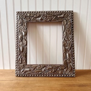 Könnte beinhalten: Ein rechteckiger, verzierter Bilderrahmen mit dunkelbraunem Finish. Der Rahmen weist detaillierte Blatt- und Blumenschnitzereien auf, die mit kleinen, silberfarbenen Nieten an den Rändern akzentuiert sind. Der Rahmen wird auf einer Holzoberfläche präsentiert.
