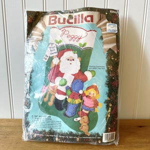 Könnte beinhalten: Ein Bucilla Weihnachtsstrumpf-Set mit einem Weihnachtsmann-Design, einem Mädchen, einem Hund und anderen festlichen Elementen. Das Set enthält den Namen "Peggy" und den Text "A Visit With Santa". Das Set ist in einer transparenten Plastiktüte verpackt.
