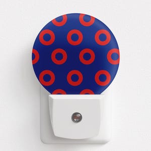 May include: A white night light with a blue and red circular pattern. The night light is plugged into a white wall outlet. The circular pattern is composed of red circles on a blue background.