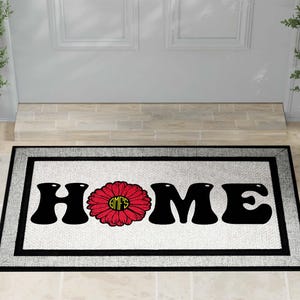 May include: A rectangular doormat with a black border and a white center. The word "HOME" is spelled out in large, black, bubble letters. A red flower with a yellow center replaces the "O".