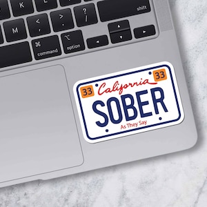 May include: A white sticker in the shape of a California license plate with the word "SOBER" in large navy blue letters. The text "As They Say" is below. The sticker is on a laptop.