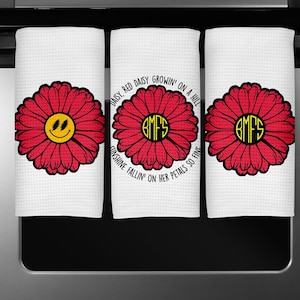 May include: Three white kitchen towels with red daisy flower designs. One daisy has a yellow smiley face, and the others have the initials "BMFS" in a yellow circle. The middle towel has the text "DAISY, RED DAISY GROWIN' ON A HILL SUNSHINE FALLIN' ON HER PETALS SO FINE."