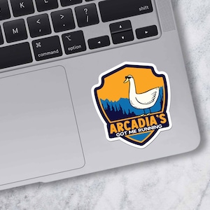 May include: A sticker featuring a white goose inside a shield-shaped design. The shield has an orange and blue background with the text "Arcadia's Got Me Running". The sticker is placed on a laptop.