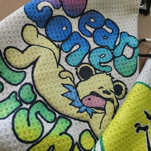 May include: A white waffle-textured towel featuring a graphic of a yellow bear with a blue collar, surrounded by the text "BEAR GONE FISHIN" in a colorful gradient of pink, blue, and green. The towel includes a black metal grommet and clip.