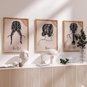 May include: Three framed art prints with line drawings of braided hairstyles. The prints are in gold frames, with the words "Beautiful", "Brave", and "Bold" below each image. The artwork is displayed above a white console table.