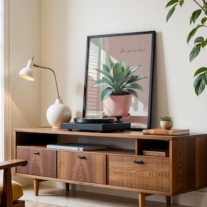 Puede incluir: Una consola de medios de madera de estilo moderno de mediados de siglo con tres cajones y un estante. Una impresión enmarcada de una planta en una maceta rosa cuelga sobre un tocadiscos. Una lámpara blanca y una pequeña planta en maceta también están en la consola.