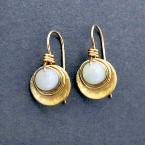 May include: A pair of gold earrings with a white stone set in a hammered gold disc. The earrings have a simple wire hook closure.