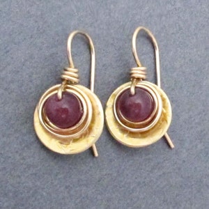 May include: A pair of gold earrings with a round, dark purple gemstone set in the center of each earring. The earrings are made with multiple layers of hammered gold wire.
