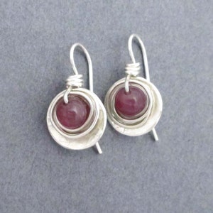 May include: A pair of silver earrings with a round, dark purple gemstone set in the center of each earring. The earrings have a layered, textured design.