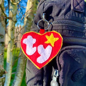 May include: A heart-shaped red felt keychain with a yellow star and the white letters "ty". The keychain is attached to a silver ring and a red ribbon. The keychain is hanging on a black backpack.