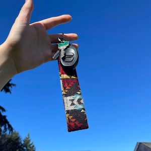 May include: A car key fob with a patterned fabric wrist strap. The strap features a geometric design in red, black, yellow, and white. The key fob is black and silver, with a key attached.