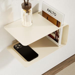 Peut inclure: Une table de chevet murale crème à deux étagères. L'étagère supérieure contient un vase en verre transparent avec des roseaux bruns et des fleurs blanches, et un livre intitulé "AALTO". Un smartphone noir repose sur l'étagère inférieure.