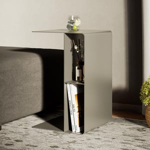 Puede incluir: Una mesa auxiliar de metal color topo en forma de C con una superficie superior plana y un compartimento de almacenamiento vertical dividido en dos estantes. El estante superior contiene un difusor de vidrio con varillas y el inferior dos libros.