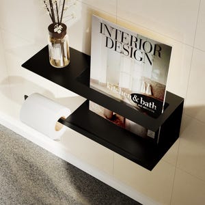 Puede incluir: Un estante de baño de metal negro con una revista, papel higiénico y una pequeña botella de vidrio con cañas. La revista dice "INTERIOR DESIGN" y "kitchen & bath". El estante está montado en una pared de azulejos color crema.