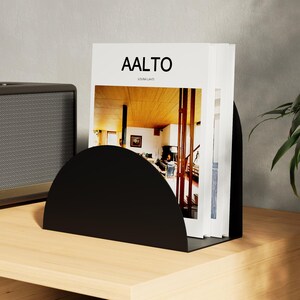 Pode incluir: Um suporte de livros de metal preto em forma de semicírculo segura uma pilha de livros com o título "AALTO" na capa. O suporte de livros está sobre uma superfície de madeira clara, ao lado de um alto-falante.