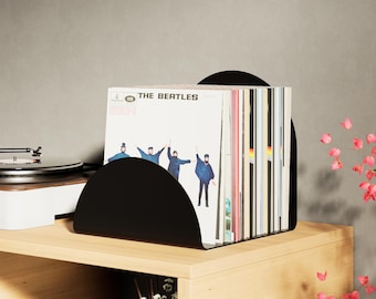 May include: A black, semi-circular vinyl record holder on a wooden shelf. The holder contains several vinyl records, including one with "THE BEATLES" printed on the cover. A record player is visible in the background.