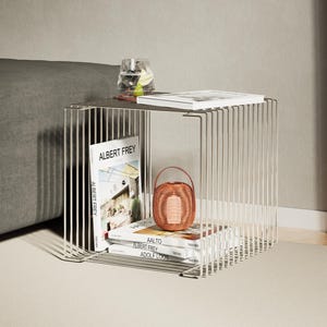 May include: A modern, open-frame side table made of thin, vertical metal rods. The table holds books, a small woven basket, and a glass of water. The books include titles like "Albert Frey" and "Aalto".