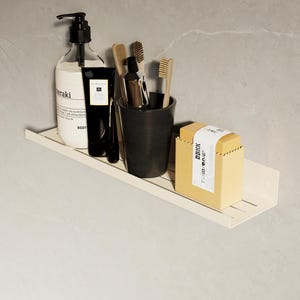 Puede incluir: Una estantería metálica de color crema con artículos de baño. Incluye un dispensador de jabón, una taza negra con cepillos de dientes, un tubo de loción y una pastilla de jabón en una caja de cartón. La estantería está montada en una pared de azulejos grises.
