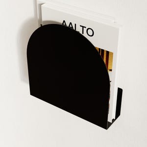 May include: A black, wall-mounted magazine holder with a semi-circular top. The holder contains several magazines, with the word "AALTO" visible on the cover. The holder is mounted on a white wall.