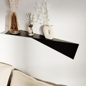Puede incluir: Estantería flotante de metal negro moderno montada en una pared blanca, que muestra objetos decorativos. La estantería contiene un jarrón de cristal marrón con flores secas, una pequeña botella de cristal con cañas y un jarrón de cerámica blanco con un diseño recortado único.