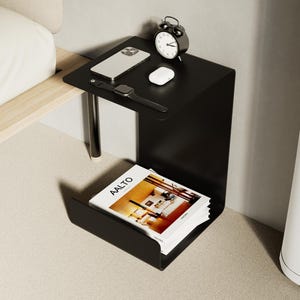 May include: A contemporary black bedside table with a minimalist design. It features a top surface with a smartphone, smartwatch, and a small white device, plus a classic alarm clock. A lower shelf holds a magazine. The table is supported by a single chrome leg.