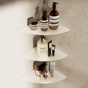 Puede incluir: Tres estantes de esquina blancos montados en una pared de azulejos grises, cada uno con varios productos cosméticos y para el cuidado de la piel. Los productos incluyen botellas, frascos y tubos con etiquetas, creando una estética minimalista de baño o spa.