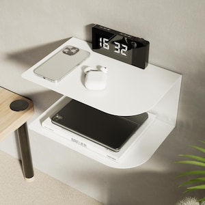 May include: A white, wall-mounted bedside table with two shelves. The top shelf holds a silver smartphone, white earbuds, and a black digital clock showing 16:32. The bottom shelf has a black tablet and a book.