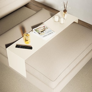 Può includere: Un pouf beige imbottito con un tavolino bianco attaccato. Il tavolo contiene una rivista, un telecomando, un bicchiere di liquido, candele e un vaso con bastoncini decorativi. Il pouf ha gambe in legno scuro.