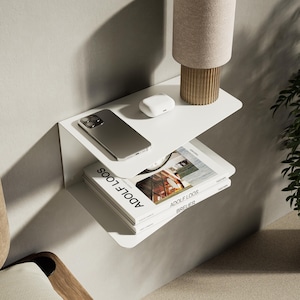 Könnte beinhalten: Eine moderne, weiße Wandregaleinheit mit zwei Ebenen. Auf der oberen Ablage befinden sich ein Smartphone, kabellose Ohrhörer und eine kleine Lampe mit beigem Schirm. Auf der unteren Ablage liegt ein Buch mit dem Titel "ADOLF LOOS". Im Hintergrund eine Farnpflanze.