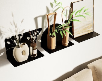 May include: Three black metal floating shelves mounted on a white wall, each holding decorative items. The shelves display a cream-colored vase, small glass bottles, bamboo containers, and a framed picture. The items are filled with plants and flowers.