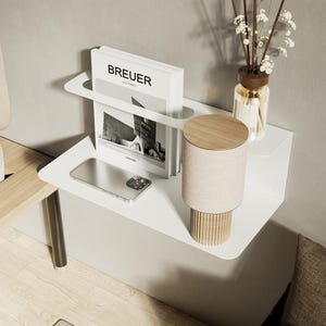 Könnte beinhalten: Ein weißes, schwebendes Nachttischregal mit einem Buch, Telefon und einer Lampe. Das Buch trägt den Titel "BREUER". Die Lampe hat einen beigefarbenen Schirm und einen Holzsockel. Eine Glasflasche mit Schilf und Blumen steht auf dem Regal.