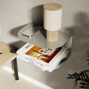 Puede incluir: Una mesita de noche moderna, montada en la pared, de metal plateado. Presenta un diseño curvo con dos estantes. Una pequeña lámpara con pantalla beige y base de madera se encuentra en el estante superior, junto con un smartphone. Los libros están apilados en el estante inferior.
