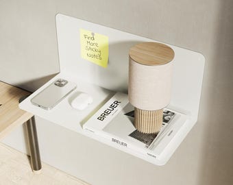 Può includere: Una mensola bianca a parete con uno smartphone, auricolari wireless e un libro intitolato "BREUER". Una lampada cilindrica con base in legno e paralume beige è appoggiata sulla mensola. Un post-it giallo con testo è attaccato al muro.