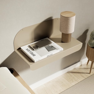 Peut inclure: Une table de chevet beige fixée au mur avec un dos incurvé. Une petite lampe avec une base en bois et un abat-jour beige est posée sur la table, ainsi qu'un livre et un téléphone. Un petit cactus en pot est visible à droite.