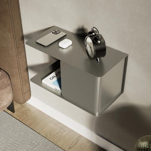 Peut inclure: Une étagère murale flottante en métal gris aux coins arrondis. La surface supérieure supporte un smartphone, des écouteurs sans fil blancs et un réveil analogique noir. Un livre avec le texte CALATRAVA est visible sur l'étagère inférieure.