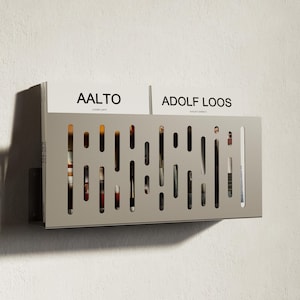 May include: A light grey metal magazine rack with a slotted design, mounted on a white wall. The rack holds magazines and books, with the titles "AALTO" and "ADOLF LOOS" visible. The rack is designed for wall mounting.
