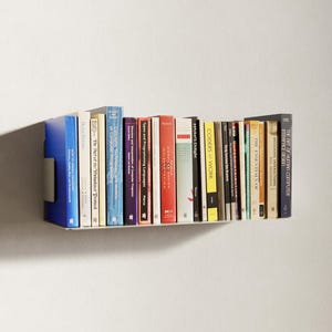 May include: A white floating shelf displaying a selection of books with colourful spines. Titles visible include "The Art of Human-Computer Interface Design" and "Coders at Work". The shelf is mounted on a white wall.